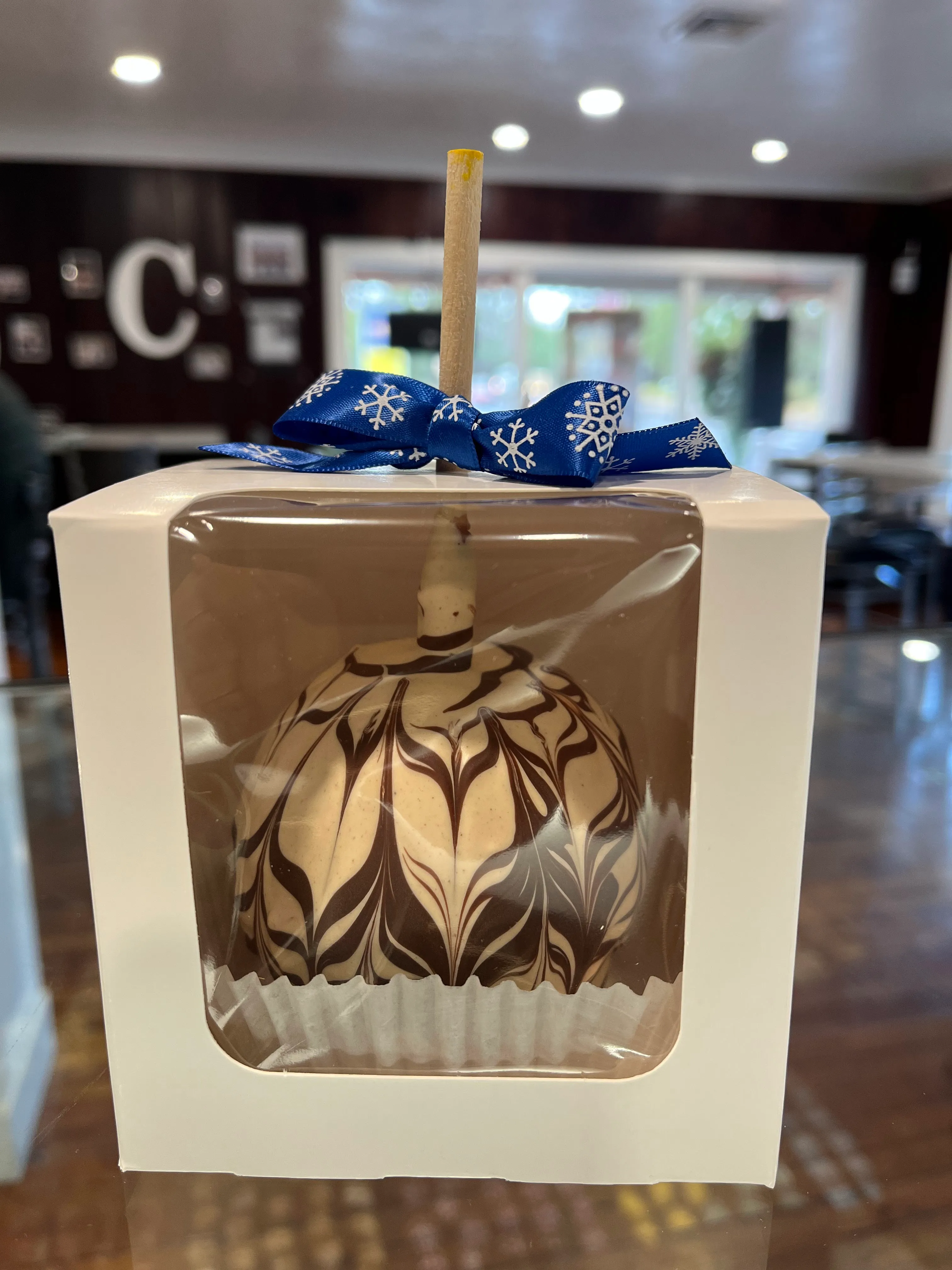 A chocolate-marbled caramel apple in a white window box with a snowflake-printed blue ribbon