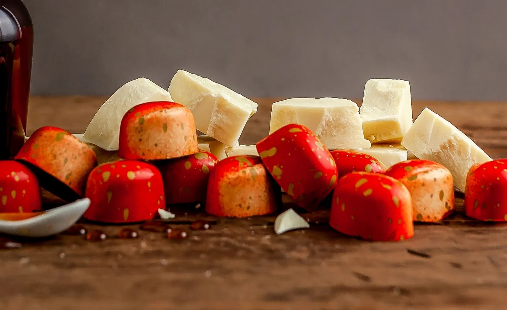 Red and orange speckled strawberry-shaped chocolates beside white chocolate chunks on a wooden surface