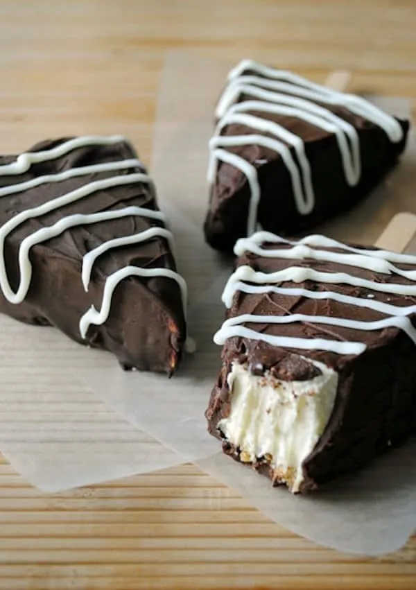 Slices of frozen cheesecake dipped in chocolate with white chocolate drizzle, on sticks