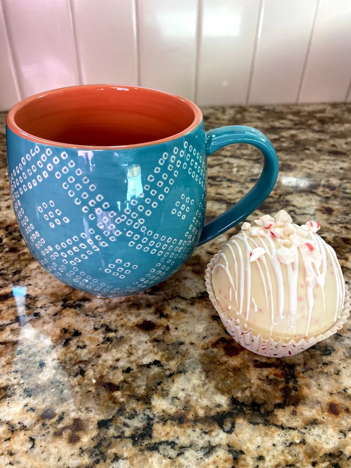 A peppermint hot chocolate bomb beside a teal patterned mug on a granite countertop